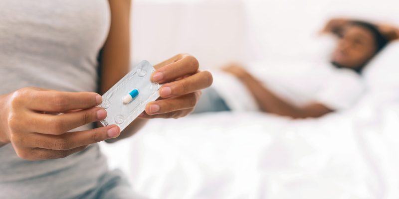 Black woman taking oral emergency contraceptive after sex Oral emergency contraception. Black woman taking medicine after sex in bed, making decision of OCP, worried about side effects, boyfriend on background